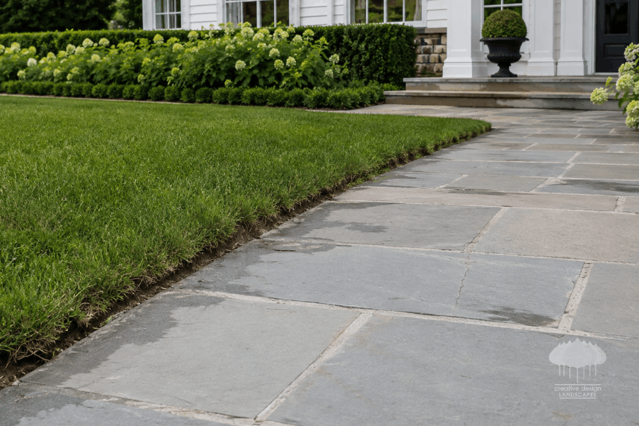 A premium Hamptons property showcases an elegant bluestone walkway and manicured lawn edge with subtle signs of seasonal wear, including softened turf lines and minor settling. The image demonstrates why a missed season is easy to see in luxury Hamptons seasonal landscaping.